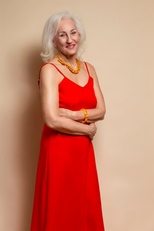 Smiling Happy Senior Caucasian Blond Woman Posing in Red Dress In Studio Over Beige Background With Positive Facial Expression. Vertical Imageの写真素材