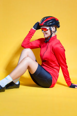 Winsome Sporty Positive Female Cyclist Sitting on Floor While Putting on Cycling Helmet Against Yellow Color Background. Vertical image Compositionの写真素材