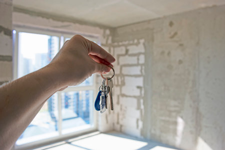 Hand of Woman Holding and Showing Keys On Fingers to Apartment Against Roughly Finished New Apartment Building Indoors. Horizontal Imageの写真素材