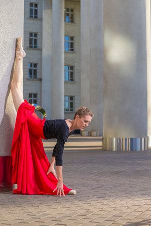 Portrait of One Young Caucasian Winsome Ballet Dancer in Black Bodysuit Posing in Red Skirt While in Dance Pose With Lifted Leg At Pillar Outdoors. Vertical Imageの写真素材