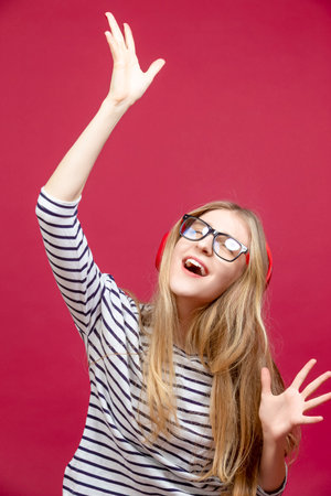 One Winsome Smiling Happy Blond Girl in Striped Casual Clothing Posing With Hand Lifted In Dance Pose Over Crimson Red Background. Vertical Shotの写真素材