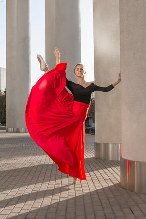 Young Caucasian Ballet Dancer in Black Bodysuit Posing in Red Skirt While in Dance Pose With Lifted Leg At Prop Against Pillars Outdoors. Vertical Shotの写真素材