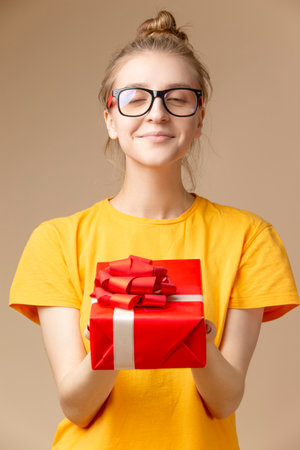 One Winsome Positive Caucasian Blond Female Girl with Red Gift Christmas Box In Round Glasses Holding And Presenting Wrapped Giftbox Against Beige. Vertical Imageの写真素材