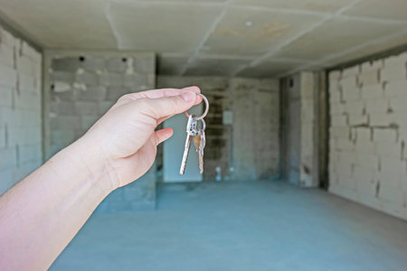 Hand of Caucasian Young Woman Holding and Showing Keys hanged On Fingers to Apartment Against Roughly Finished New Apartment Building.Horizontal Orientationの写真素材