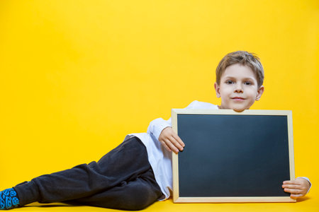 Portrait of Adorable Preschooler Caucasian Boy Holding Wooden School Blackboard While Lying On Floor With Lifted Hands Isolated On Yellow Background. Horizontal Shotの写真素材
