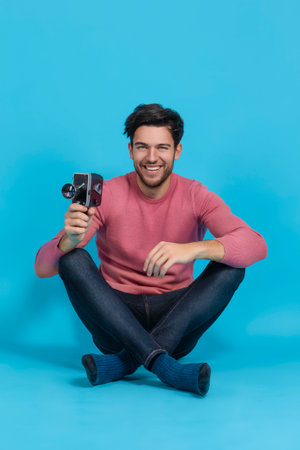 Smiling Brunet Man in Pink Coral Jumper And Hipster Glasses While Holding Retro Film Camera in Hands Sitting Against Blue Background To Depict Retro Filming Concept. Vertical Shotの写真素材