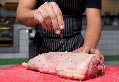 A fresh organic leg of lamb being seasoned with salt and pepper on a red chopping board.の写真素材