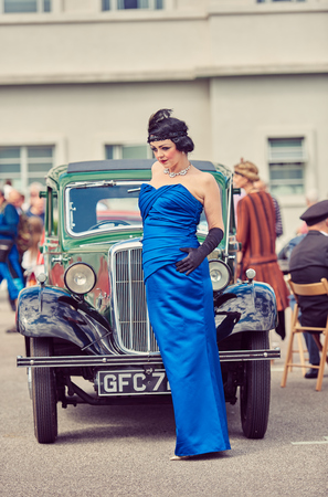 london, england, 02/05/217, A gatsby girl in 1920s Art deco style party girl flapper clothing, and feather head wear, black gloves and blue dress,  posing by a  vintage retro car. のeditorial素材