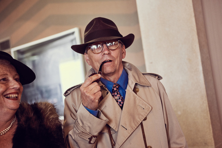 london, England, 05/05/2017, A stylish retro vintage fashionable man in a trench coat macintosh, hat and glasses smoking a pipe. Old american film noir detective inspector style clothing. Best dressed award のeditorial素材