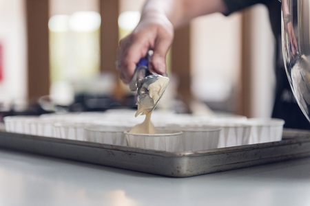 A hand holding a scoop full of creamy cake dough and putting a dollop into an empty tray of cupcake...