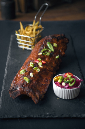 A beautiful delicious golden rack of meaty bbq pork ribs, french fries in a metal frying basket, , and chilli and salad, served on a fashionable grey slate in a dark photography environment.の写真素材