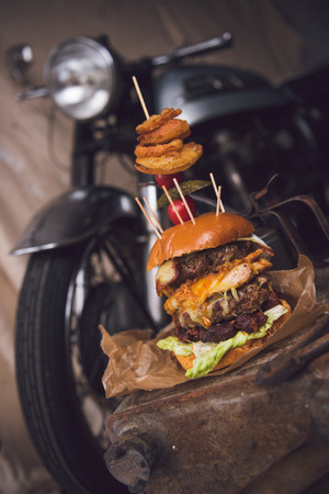 A huge stacked double cheeseburger, with bacon, cheese, lettuce, topped with gherkins, onion rings and cherry tomato. photographed in a rustic mechanic workshop background.の写真素材