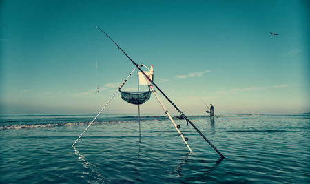 A beautiful cinematic scene of a fisherman fishing off the beach with a line and pole , with a dark blue aqua sky. Photography film colour grade.の写真素材