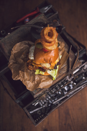 A huge stacked double cheeseburger, with bacon, cheese, lettuce, topped with gherkins, onion rings and cherry tomato. photographed in a rustic mechanic workshop background.の写真素材