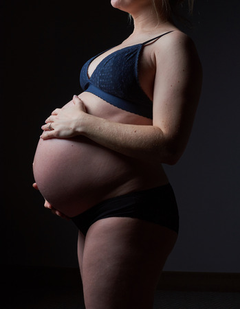 A beautiful pregnant woman body, with the woman gently holding and supporting the pregnant stomach. Gentle feminine motherhood. Photographed on a black studio background.の写真素材