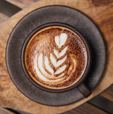 A beautiful creamy brown warm fresh cappuccino coffee cup, full of creamy coffee, served in a wooden rustic brown coffee shop with coffee art.の写真素材