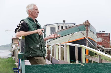 lancashire, england, 14/04/2019 A local old traditional european fisherman, standing by his boat that is moored up on a jetty by a river. Local seaman fishing industry.のeditorial素材
