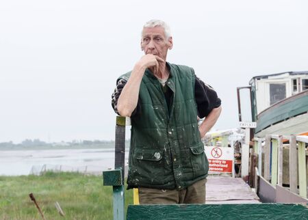 lancashire, england, 14/04/2019 A local old traditional european fisherman, standing by his boat that is moored up on a jetty by a river. Local seaman fishing industry.のeditorial素材
