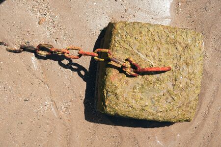 A square concrete anchor point on a sandy beach in summer. A rusty chain is attached to the home made anchorage device. Fishing boats mooring up in the water at high tide.の写真素材
