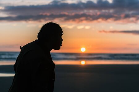 A silhouette of black male man with dreadlocks on a sunny beach with a sunset. Thinking and clarity in the great outdoors. Walking and fresh air for good positive mental healthの写真素材