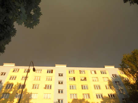 Image of large block of flats illuminated by evening sunshine, but with dark cloud above itself. Time after storm in city.の写真素材