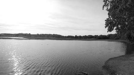 Greyscaled image - Beautiful nature scene with peaceful pond with calm water under partly cloudy sky and shining sun causing light damage due to overexposition.の写真素材