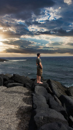 Man standing on a rock by the ocean and looking at the sunsetの写真素材
