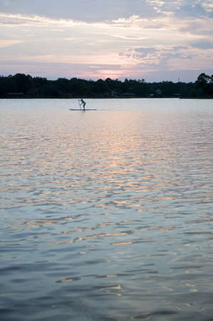 Surfer standing upright on a surboard paddling across a calm sea using a paddle against a delicate pink sunsetの写真素材