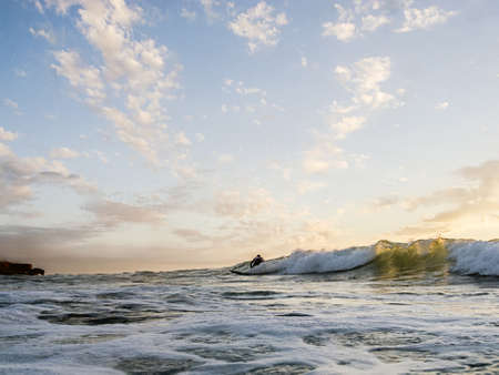 Breaking wave rolling across the ocean at sunset with a surfer silhouetted against the water in the distanceの写真素材