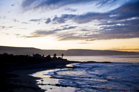 Delicate orange sunset over a coastal bay with a surfer carrying a board across the beach and a silhouetted headlandの写真素材