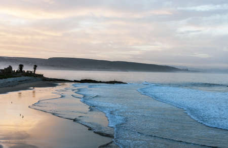 Beautiful soft glowing colours of a sunset reflecting of wet beach sand in a tranquil bay with a silhouetted headlandの写真素材