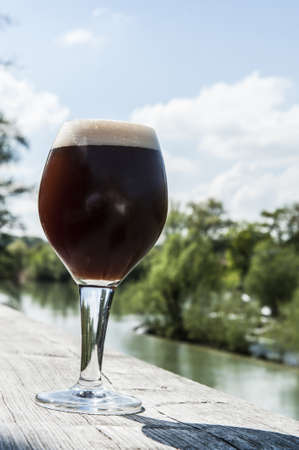 A glass of balck beer on a table beside the lakeの写真素材