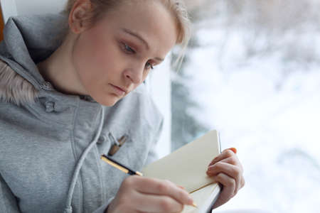 Young girl writing in her journal while sitting at a large window 