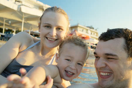 Happy young family at the seaside with a smiling attractive mother and father playing with their cute young son in the water in front of a resort complexの写真素材