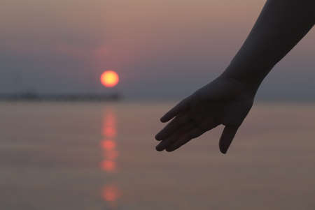 Womans hand silhouetted against an ocean sunset as the glowing orb of the sun sinks below the...