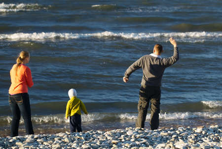 Mother, father and their little son sitting on pebble beach. Boy playing, mother and father watching moving waves, dad throwing stones into waterの写真素材
