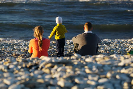 Mother, father and their little son sitting on pebble beach watching moving waves