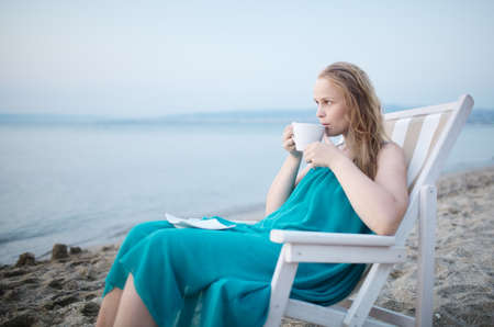 Wide shot of young girl enjoying a cup of tea at the seaside sitting relaxing on a deckchair with a blissful expression overlooking a tropical beachの写真素材