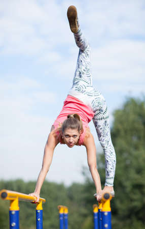 Agile attractive young female gymnast balancing on brightly coloured cross bars outdoors in a park...