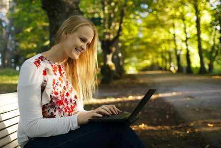 Young woman sitting on a wooden bench using her laptop in the park in a shady leafy tree lined avenueの写真素材