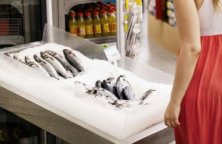 Woman shopping for fish in a supermarket standing looking at a display of fresh whole fish on iceの写真素材