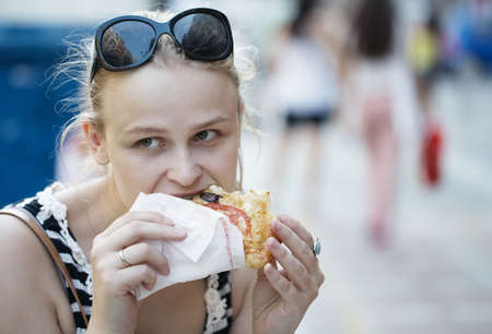 Young woman with sunglasses on her head eating fast food in the street and looking around being...