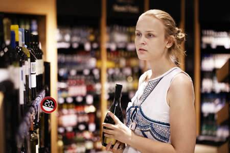Woman shopping for wine or other alcohol in a bottle store standing in front of shelves full of bottles with a serious expression as she tries to make up her mindの写真素材