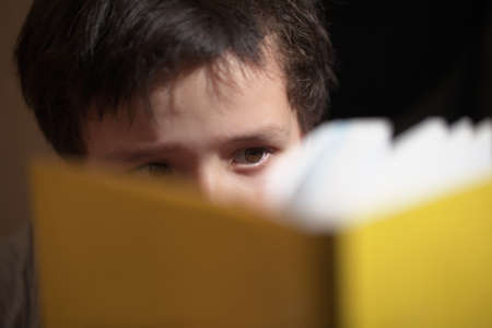 Young boy reading a yellow book with the pages concealing his face and just his eyes visible with...