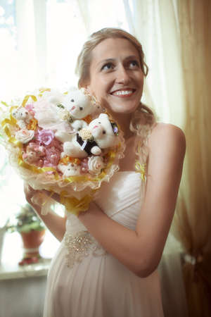 Beautiful young bride with a wedding bouquet featuring a bride and groom amongst the flowers standing in front of a windows smiling as she look up to the rightの写真素材
