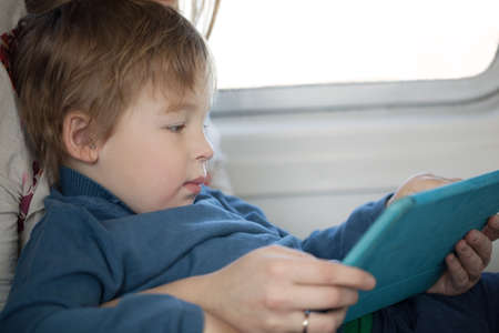Small boy sitting on his mothers lap alongside a window looking at a tablet in an airplane as he travels on vacation, side view candid portraitの写真素材