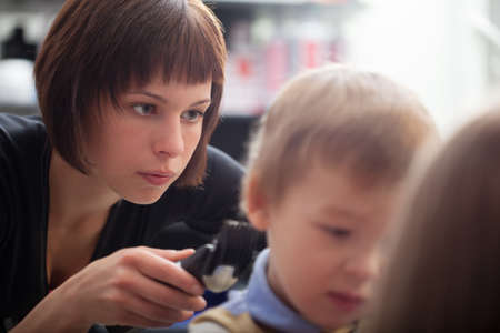 Hairstylist cutting a young boys hair bending down as she concentrates on trimming the back with a clipper, closeup portrait with focus to her faceの写真素材