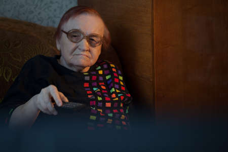 Elderly woman in glasses sitting on the sofa and changing channels on TV with remote control