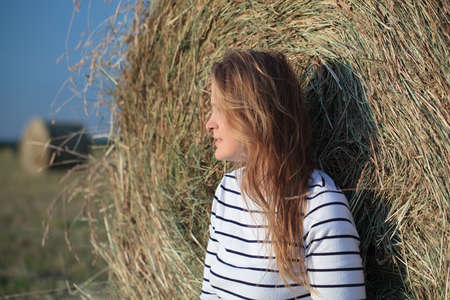 Young woman looking into the distance leaning on hay roll in the fieldの写真素材