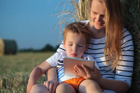 Happy mother with little son on the lap holding a pad and boy playing on it  They sitting by the hay roll, blurry field and sky in backgroundの写真素材